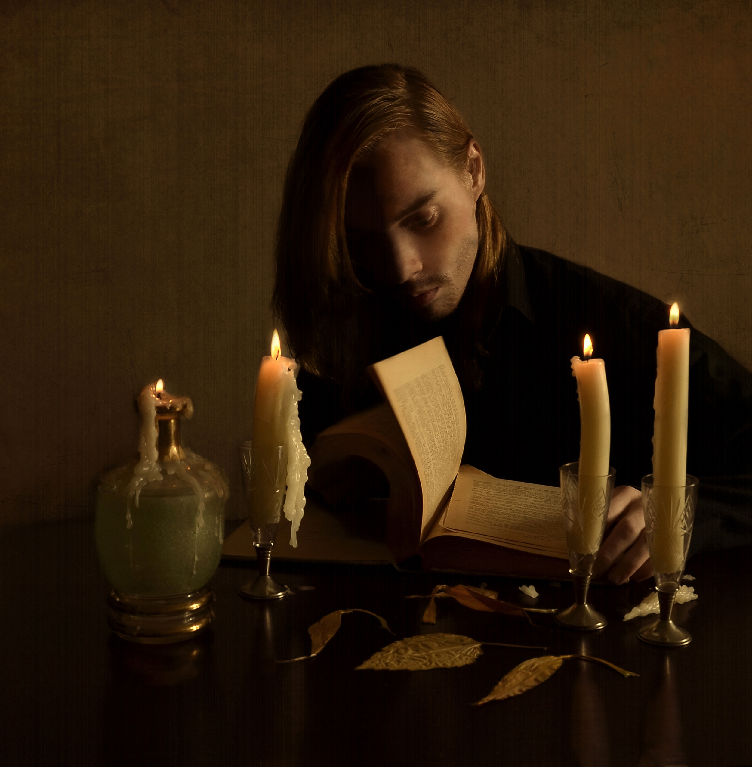 long-haired man sits at a table by candlelight over an old book and leafs through the yellowed pages